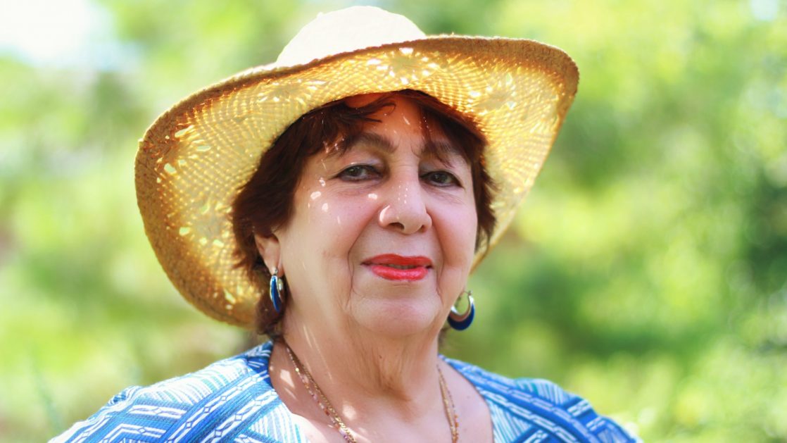 A proud older woman standing outside on a sunny day, wearing a sun hat. Green trees behind her.