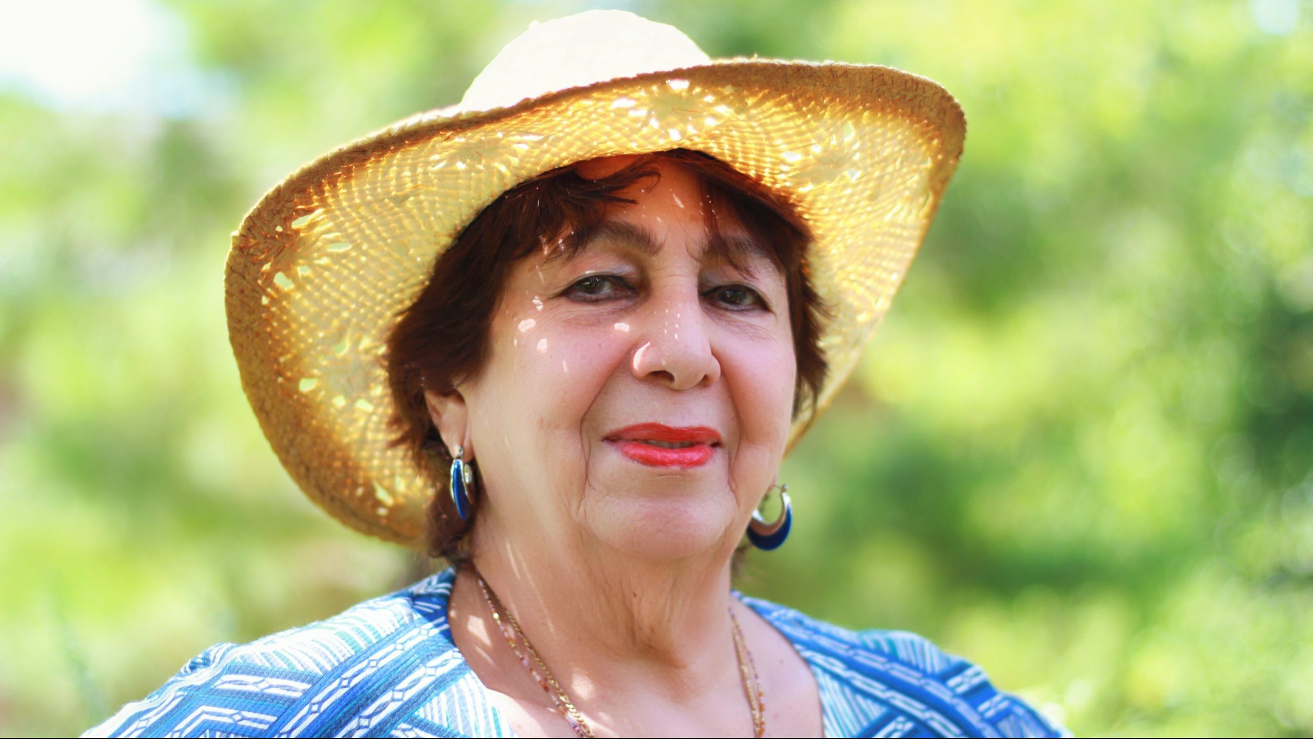 A proud older woman standing outside on a sunny day, wearing a sun hat. Green trees behind her.